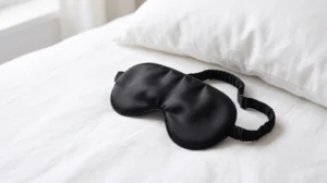 Soft black mulberry silk sleep mask on a clean white linen surface, lifestyle product shot of premium eye mask with adjustable strap, minimal composition